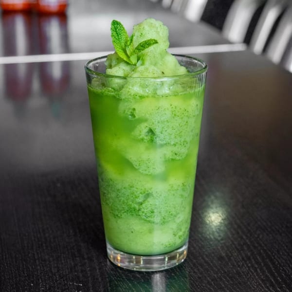A tall glass of vibrant green mint slushie, garnished with fresh mint leaves, on a dark table.