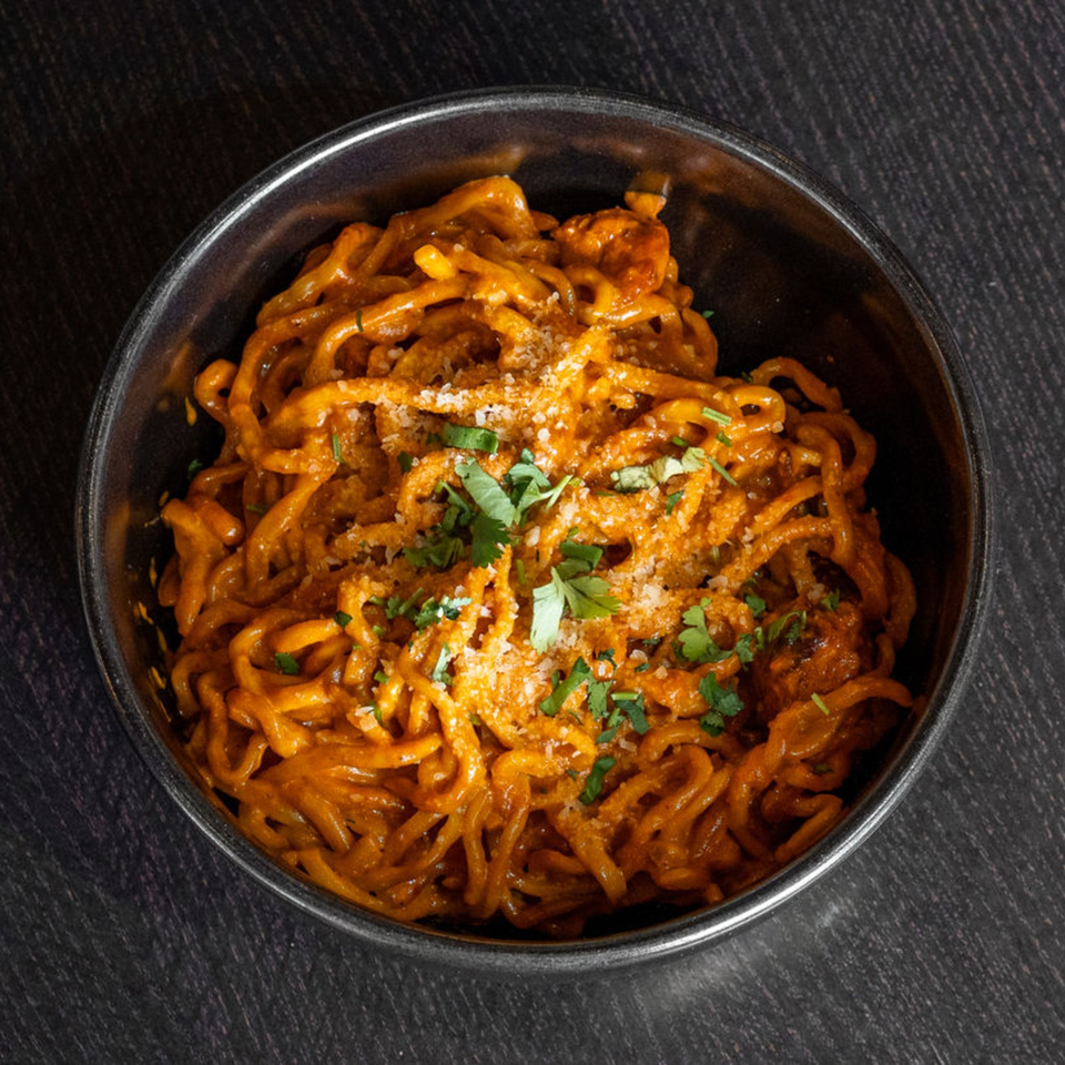 Bowl of orange-sauced noodles topped with crispy pieces, grated cheese, and fresh cilantro on a dark tabletop.