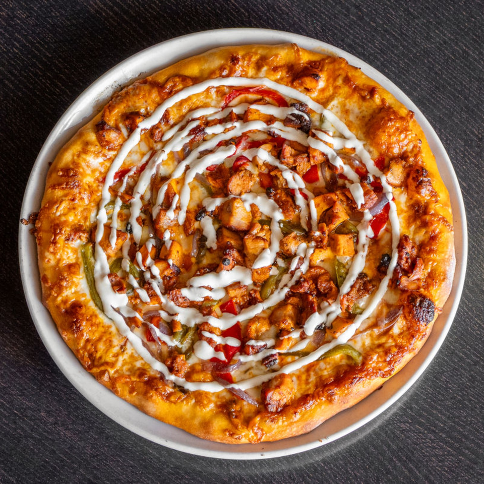 Round golden pizza topped with grilled chicken, peppers, onions, and a spiral drizzle of white sauce on a metal plate.