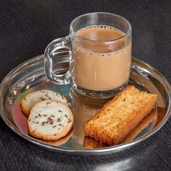 A glass mug of Mirchi Cafe chai tea, two cumin cookies, and a rusk on a silver tray.
