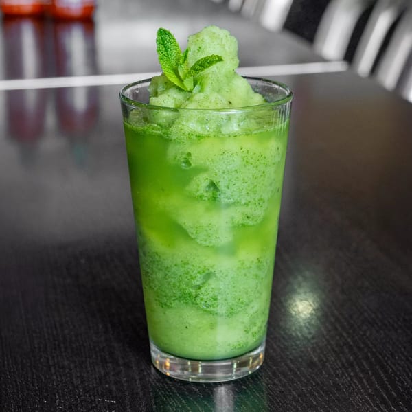 A vibrant green mint slushie in a clear glass, garnished with fresh mint leaves on a dark table.
